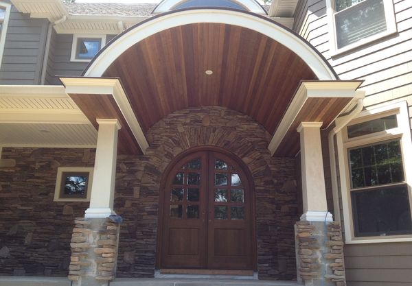Arched double wood entry doors with glass panes, set in a stone arch under a curved wood-plank porch ceiling with columns on stone bases, installed by David, who also built the porch ceiling, columns and soffits